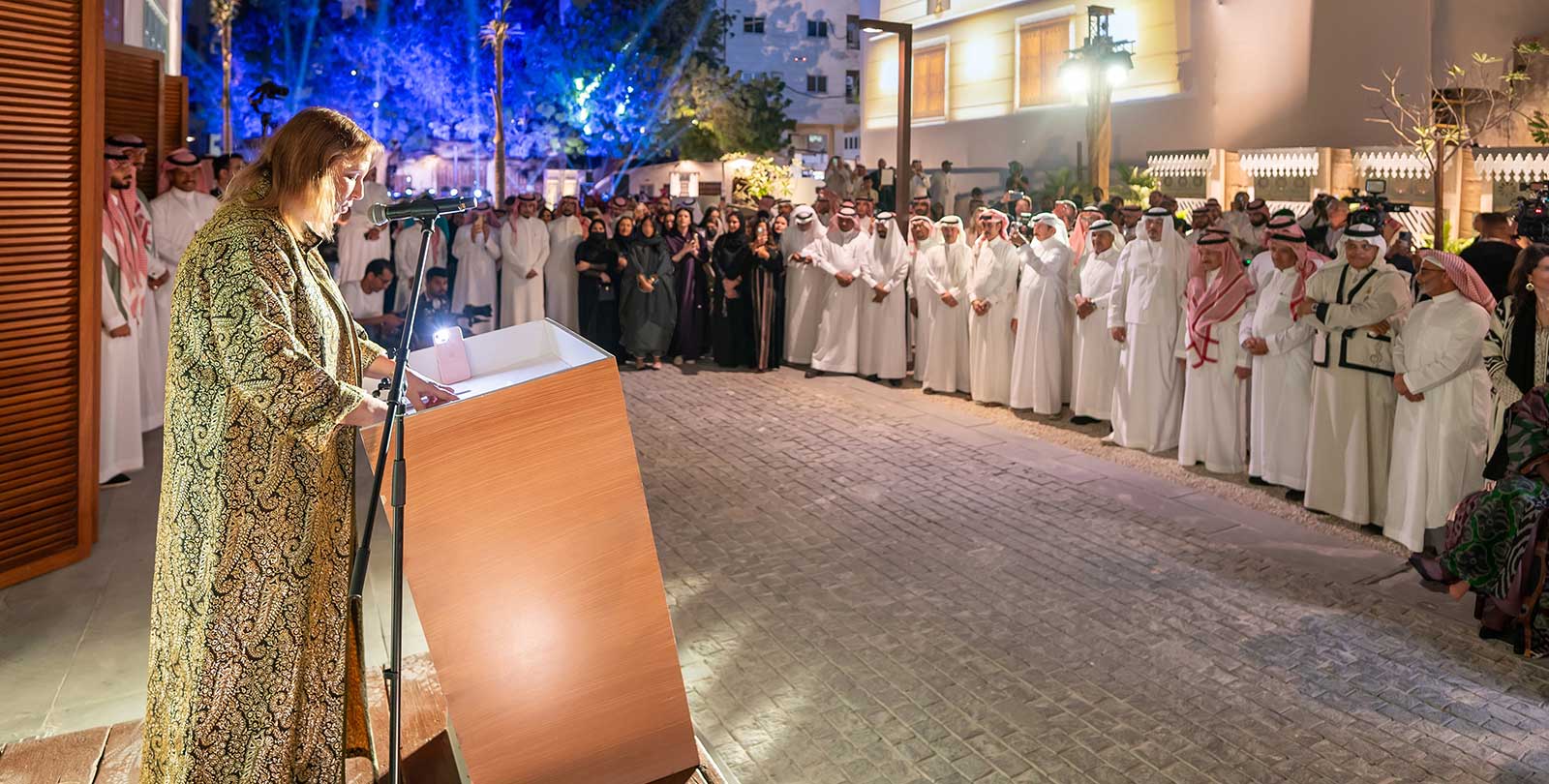 Dr. Lisa Urkevich speaking at a podium with a microphone, facing a large audience of Saudi men and women in traditional dress at an evening cultural gathering in 2024.