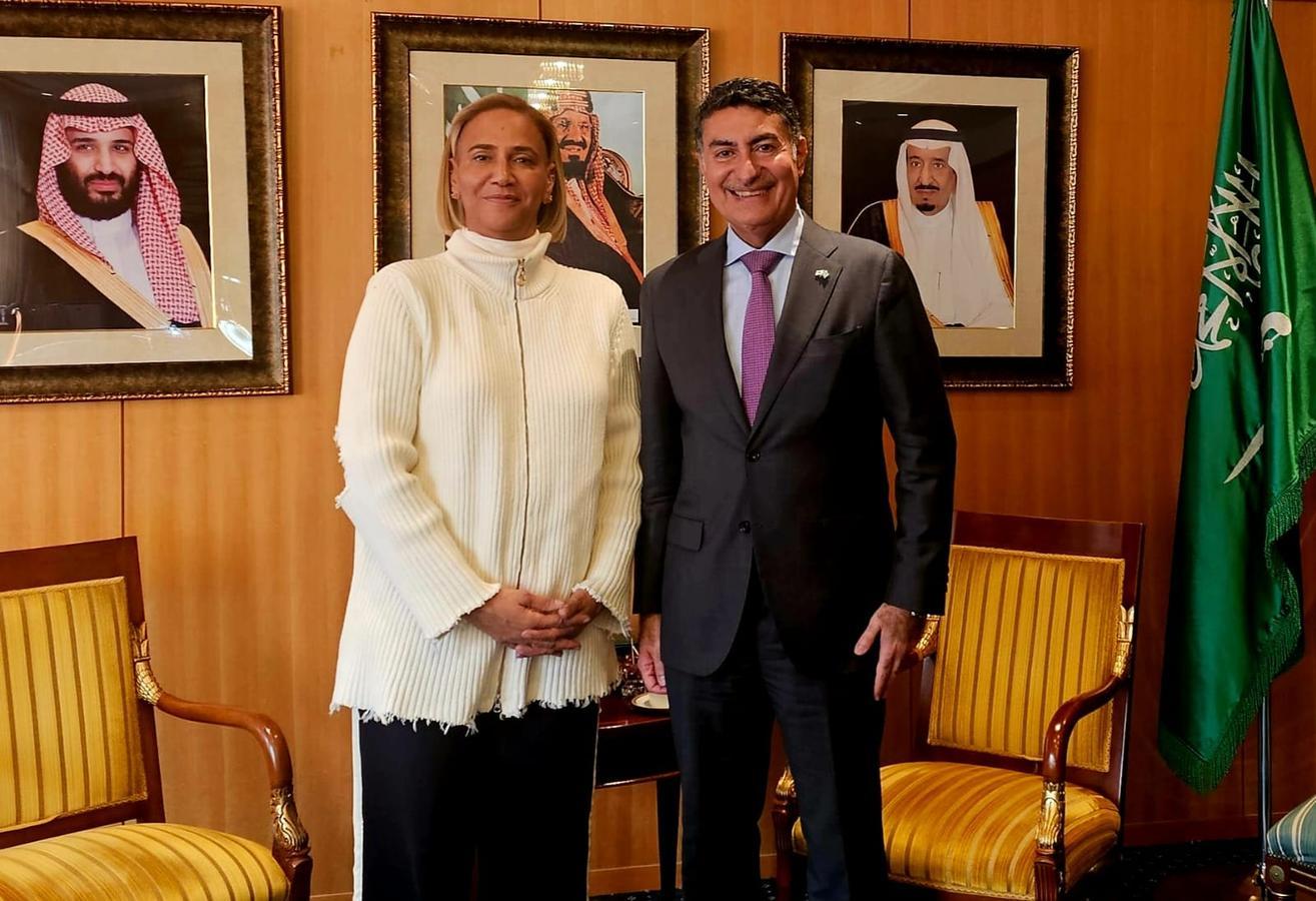 Dahlia Rahaimy stands beside Saudi Arabia’s Ambassador to Japan, Dr. Ghazi Binzagr, in front of portraits of Saudi leaders and the Saudi flag at the Saudi Embassy in Tokyo. Both are smiling, framed by elegant chairs and wood-paneled walls.