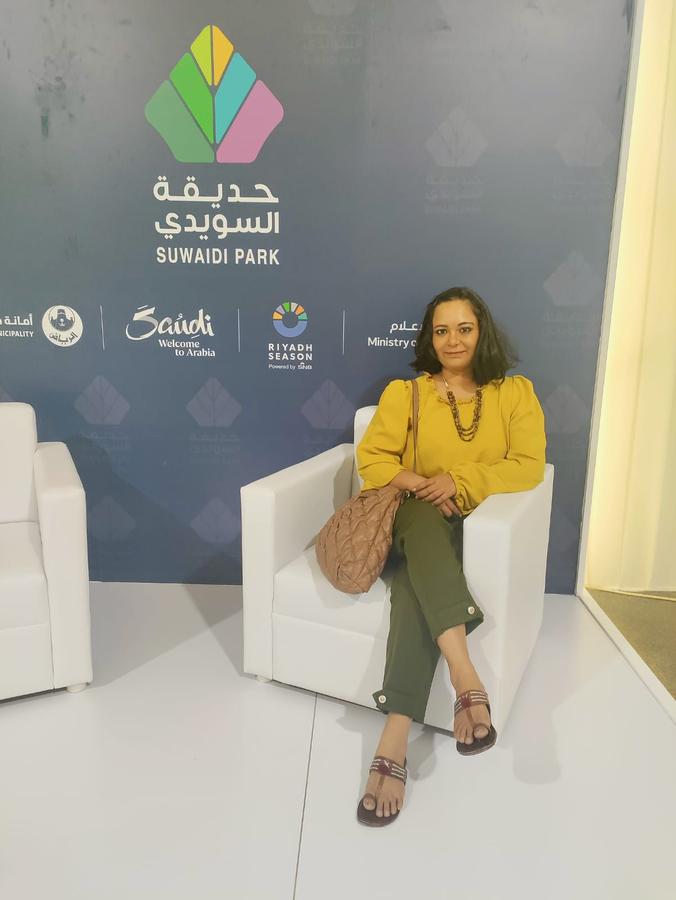 Shubhda Chaudhary seated at Suwaidi Park during Riyadh Season, wearing a mustard yellow blouse and olive green pants, smiling at the camera in front of a branded backdrop featuring the park and event logos.