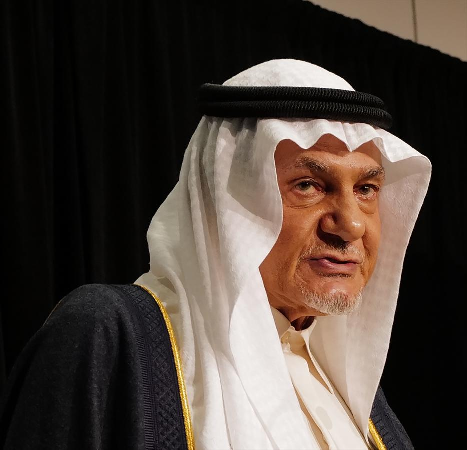 Prince Turki AlFaisal speaking at a public event, wearing traditional Saudi attire and addressing regional political developments.