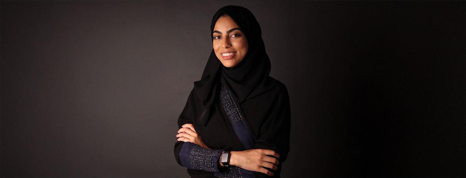 Rowa Talal Fitaihi, Saudi humanitarian and UNHCR officer, photographed against a dark background wearing a black abaya and smiling confidently.