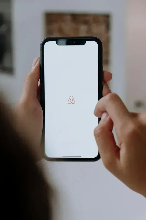 Close-up of a person holding a smartphone displaying the Airbnb logo on a white screen, with their hand interacting with the device.