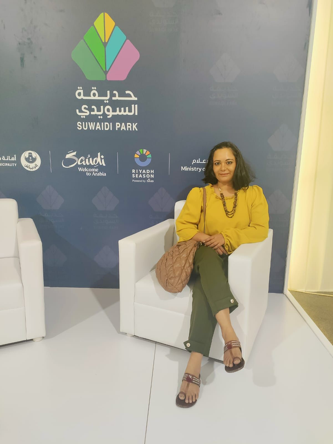 Shubhda Chaudhary seated at Suwaidi Park during Riyadh Season, wearing a mustard yellow blouse and olive green pants, smiling at the camera in front of a branded backdrop featuring the park and event logos.