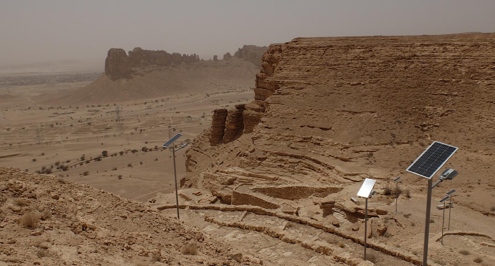 A rugged desert landscape in Saudi Arabia featuring towering cliffs and a winding stone pathway lined with solar-powered streetlights. The scene highlights the integration of renewable energy solutions in remote natural areas.