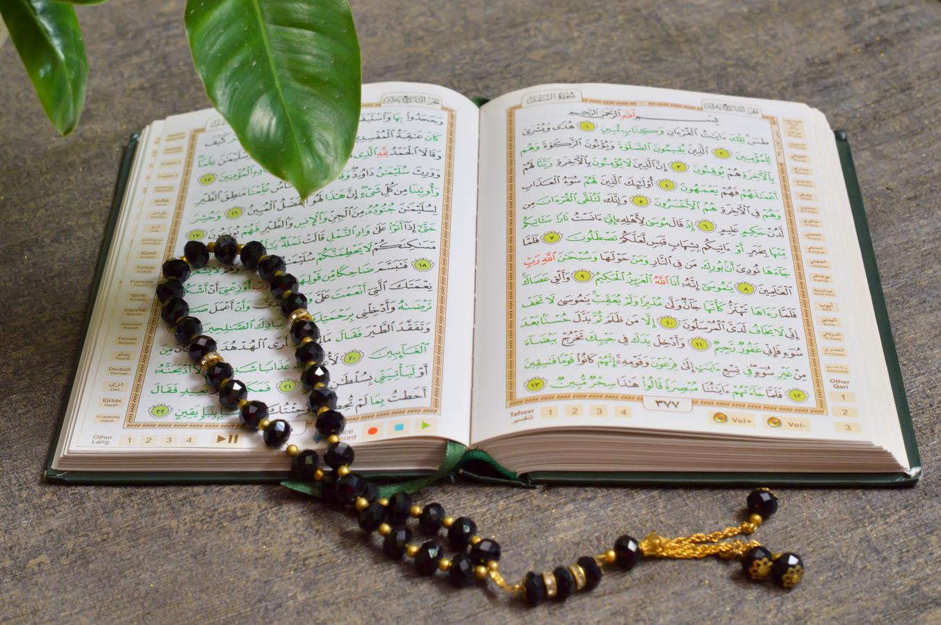 Open Qur’an with black prayer beads placed on its pages, photographed on a wooden surface with a green leaf above, representing spirituality and contemplation.