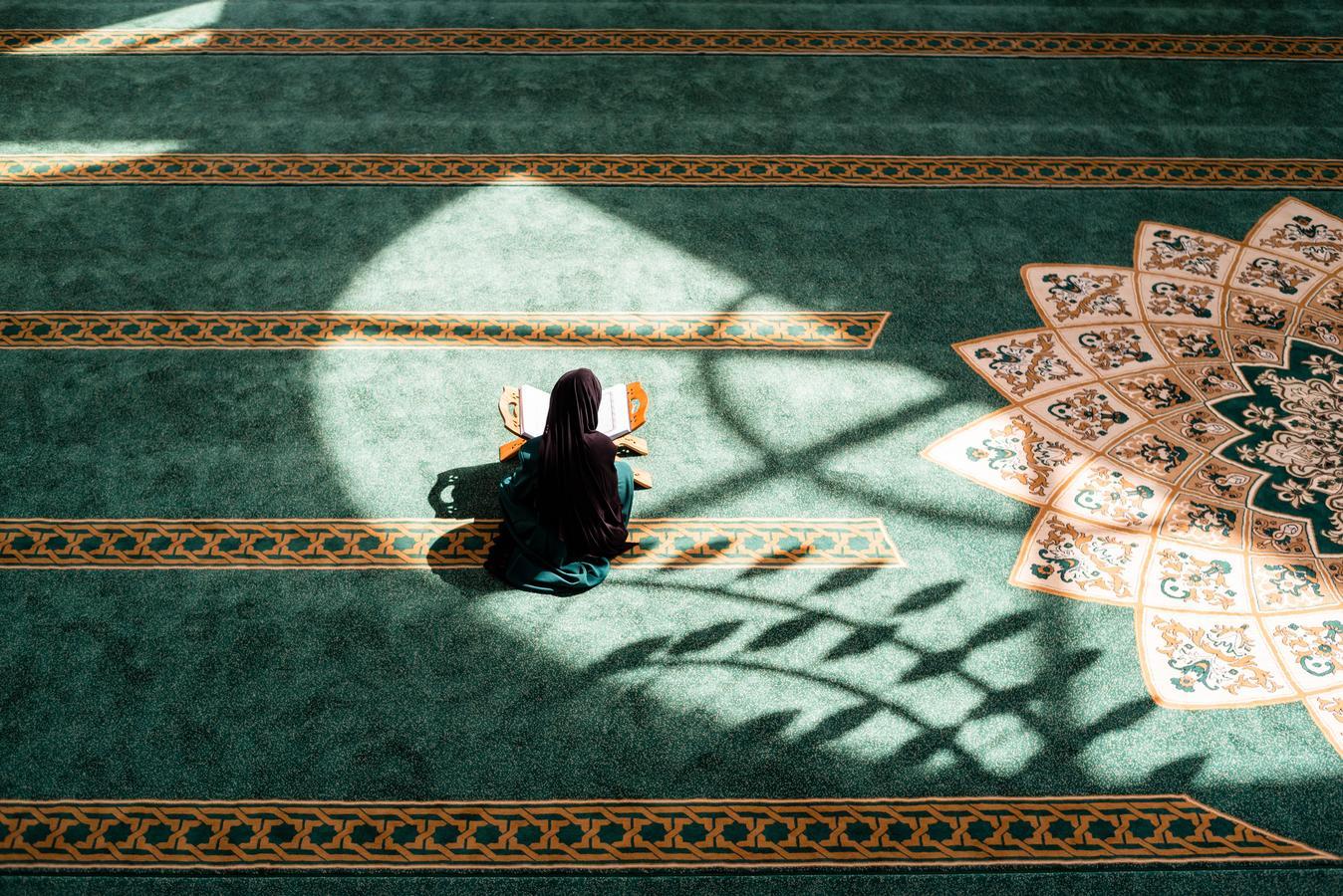 Muslim woman in hijab reading the Qur’an alone inside a mosque, sitting on green patterned carpet with sunlight and shadows.