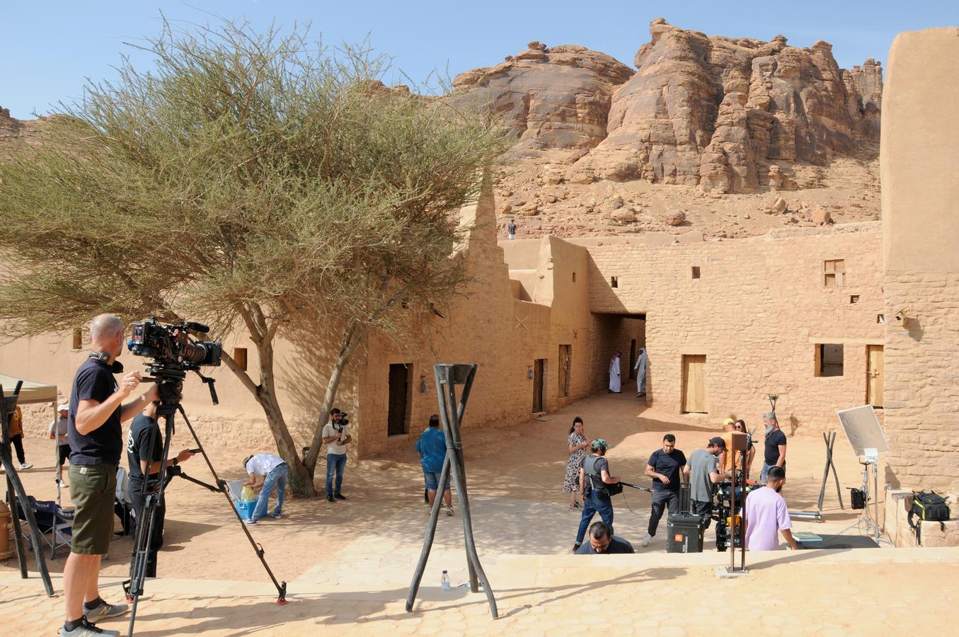A film crew sets up equipment in AlUla, Saudi Arabia, surrounded by traditional mudbrick buildings and dramatic rock formations. The scene highlights the Kingdom's growing film industry and its use of historic landscapes as cinematic backdrops.