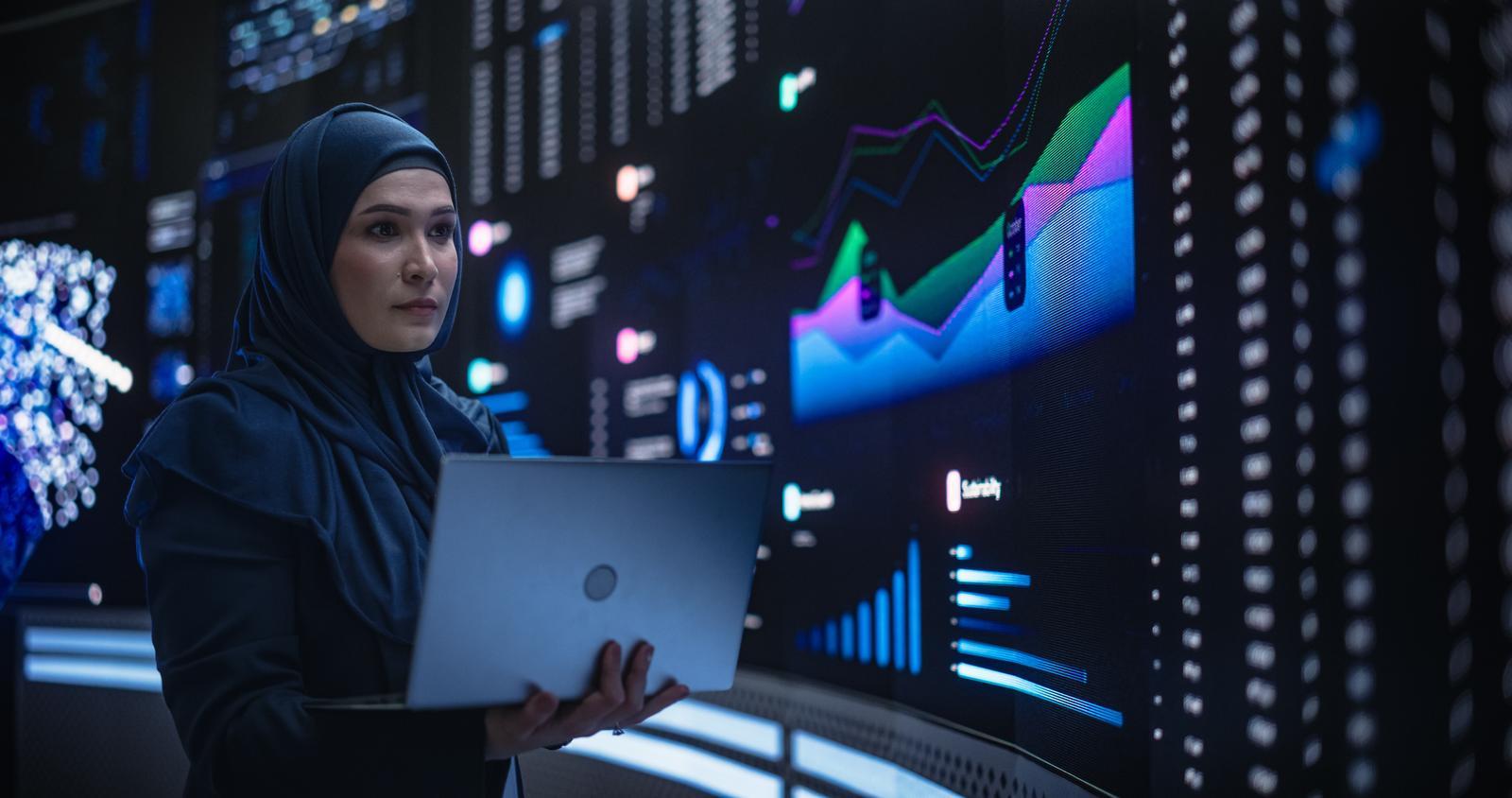 A woman standing with a laptop in a high-tech digital room, symbolizing Saudi Arabia's investment in its youth to drive progress and secure the nation's future.