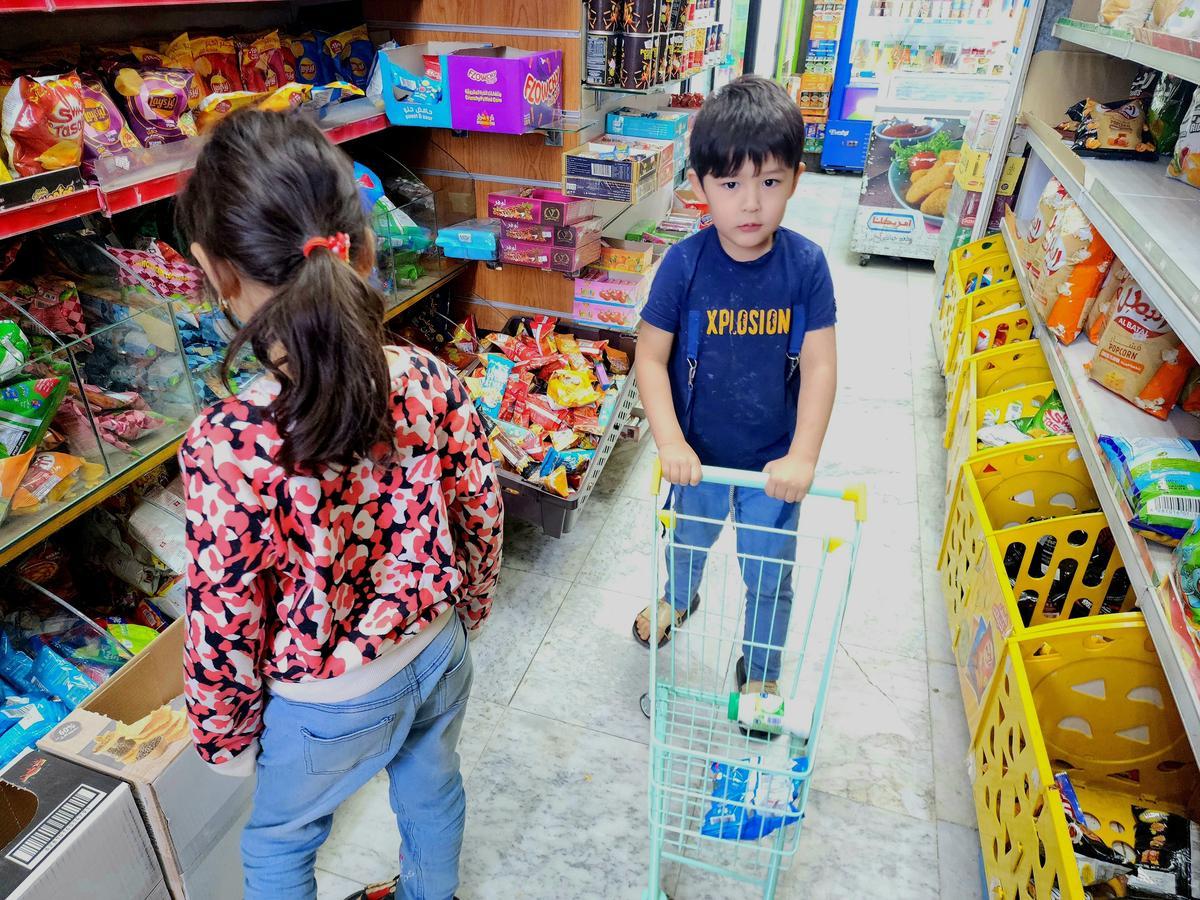 Two children inside a Saudi baqala (corner shop), one pushing a small shopping cart and the other browsing shelves filled with snacks and groceries.