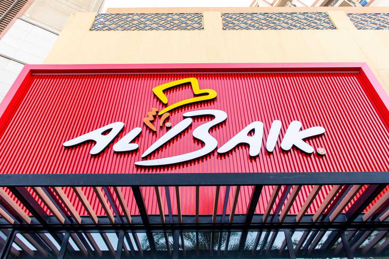 red Al Baik restaurant sign with its distinctive logo, featuring a yellow hat and stylized white lettering, displayed on the facade of a building.
