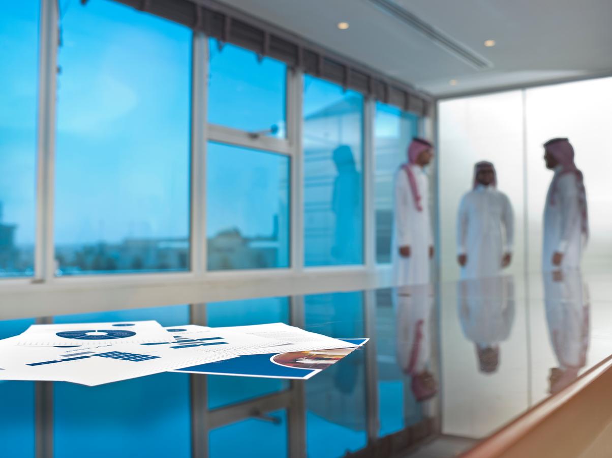 Printed business charts on a reflective office table with three Saudi men in traditional attire having a discussion in the background, symbolizing modern work culture in Saudi Arabia.