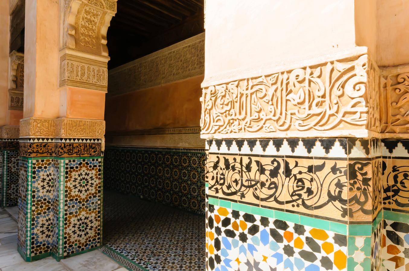 Close-up of ornate Arabic calligraphy and geometric mosaic tilework on stone columns outside a historic building in Marrakech, Morocco.