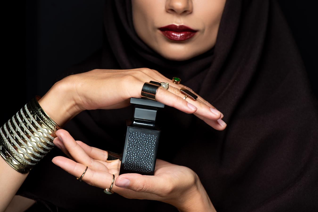 A woman in a dark hijab, wearing gold and gemstone jewelry, holds a black perfume bottle. The image highlights the intersection of modern luxury and traditional style.