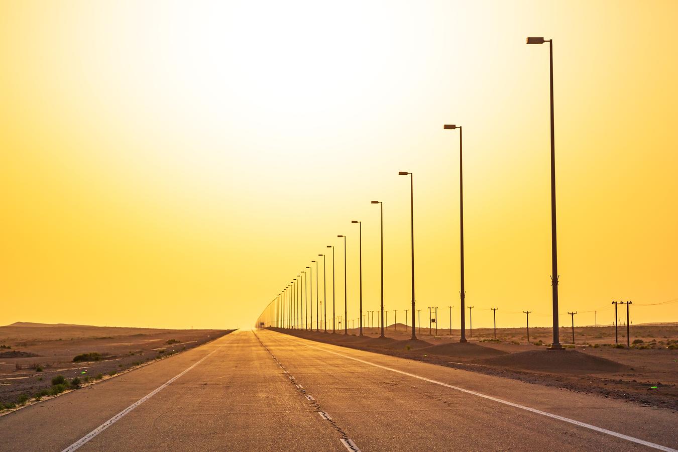 A deserted highway in Saudi Arabia under the intense midday sun, with heat shimmering over the asphalt and a long line of streetlamps stretching into the horizon.