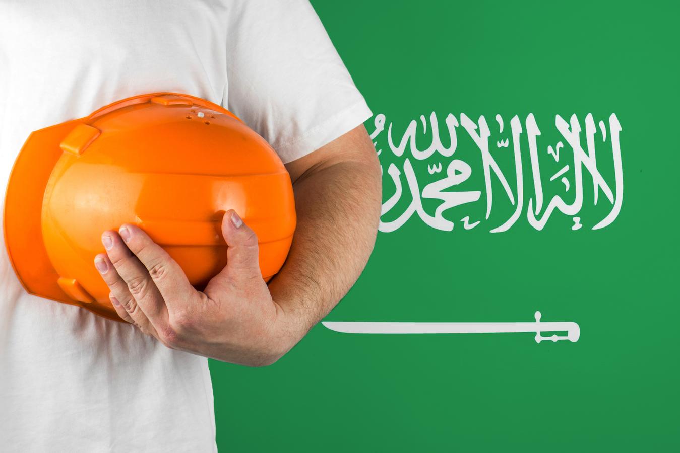 A person wearing a white shirt holds an orange construction helmet in front of the Saudi Arabian flag, representing Saudization and the rise of local employment in Saudi Arabia’s labor market.
