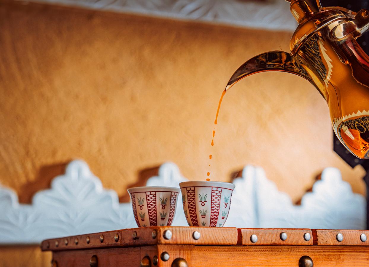 Golden dallah pouring Arabic coffee into a decorated finjan cup, with another cup beside it on a wooden table against a warm-toned traditional backdrop