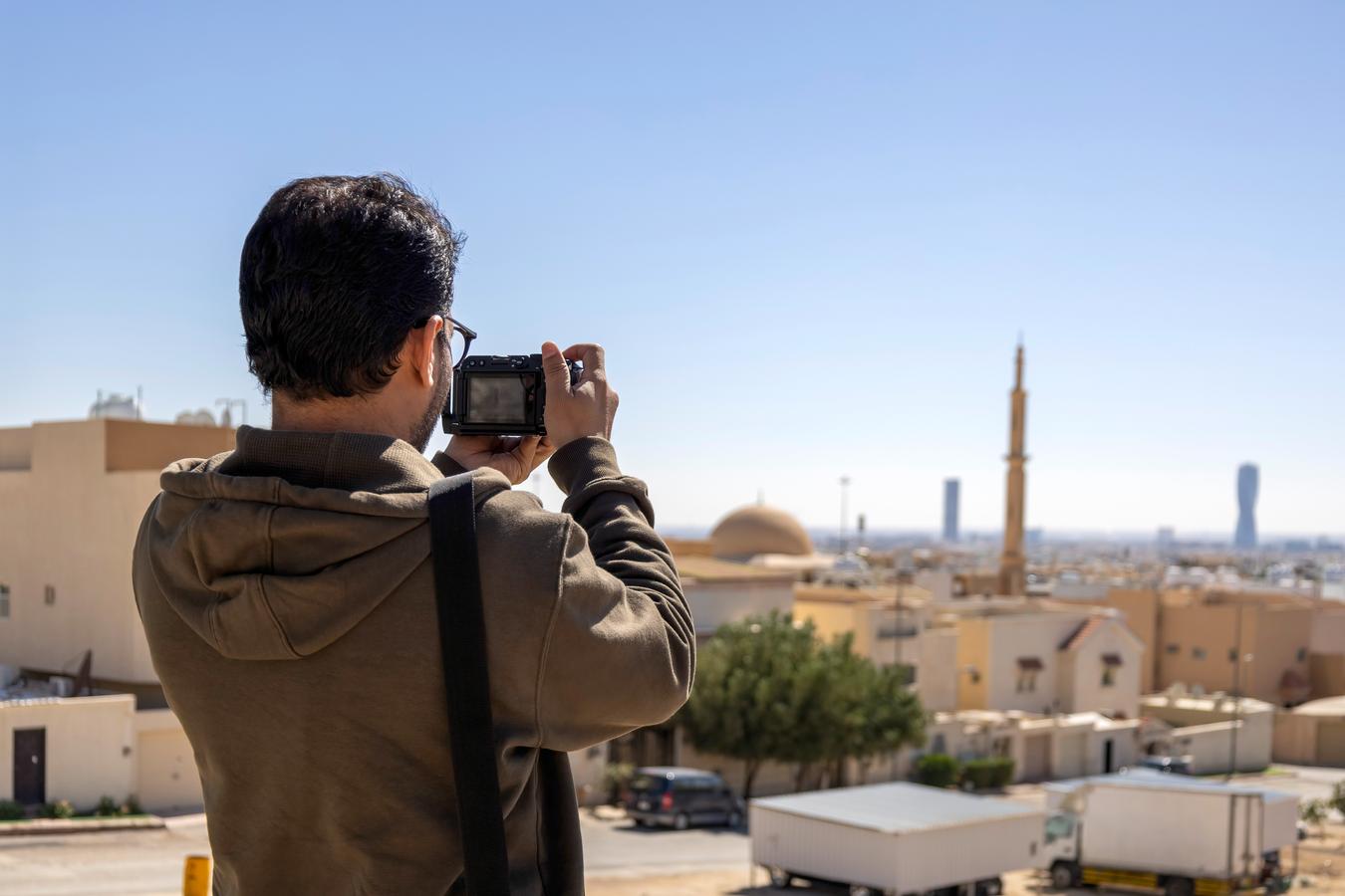 Tourist taking a photo in a residential area of Riyadh with the city skyline and mosque minaret in the background, illustrating photography rules in Saudi Arabia.