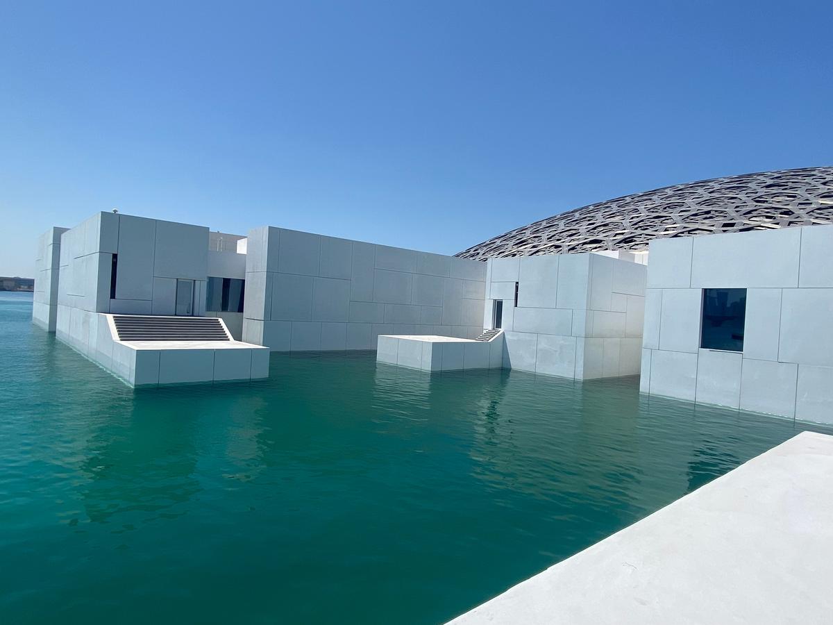 View of the Louvre Abu Dhabi’s white geometric buildings set over turquoise water, with the museum’s dome behind them, seen on a clear Gulf morning.